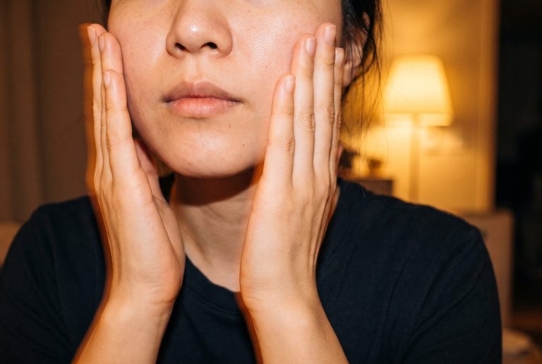 A woman presses both palms to her cheeks to pat in skincare, her face cropped below the eyes