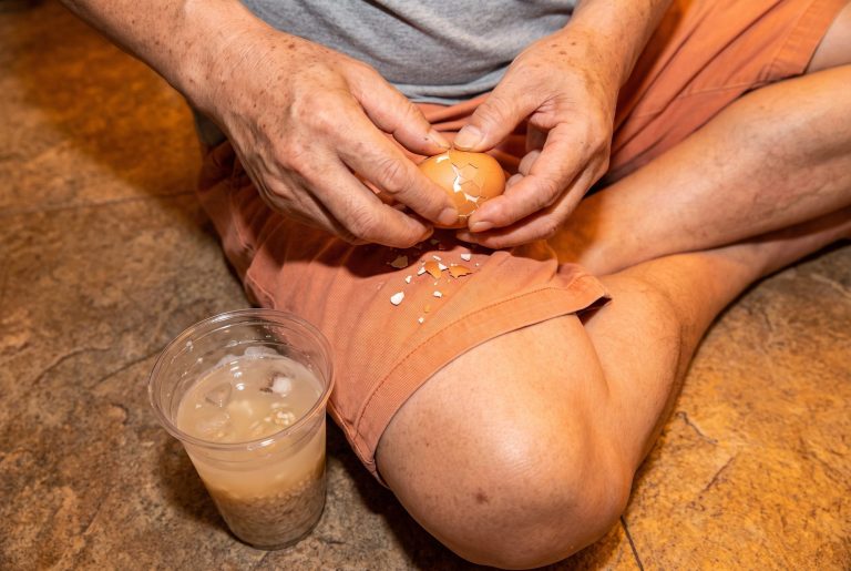 Hands peel a brown baked egg over orange bathhouse shorts beside a cup of sweet rice drink