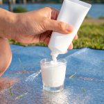 A hand squeezes white sunscreen from a plain tube into a small shot glass on a picnic mat by a river