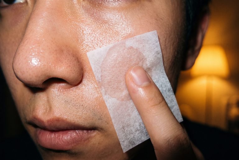 A fingertip presses a blotting paper to an oily cheek beside the nose, the paper turning see-through