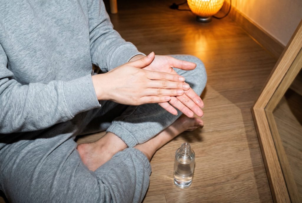 A woman in a grey sweatsuit sits cross-legged on a wooden floor, pressing toner between her palms