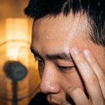 A young man pats a sheer gel sunscreen onto his shiny forehead on a humid night, a fan behind him