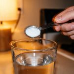 A hand holds a measuring spoon of fine white powder above a glass of water