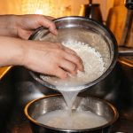 Hands rinse short-grain rice in a steel bowl, the milky water pouring into a second bowl